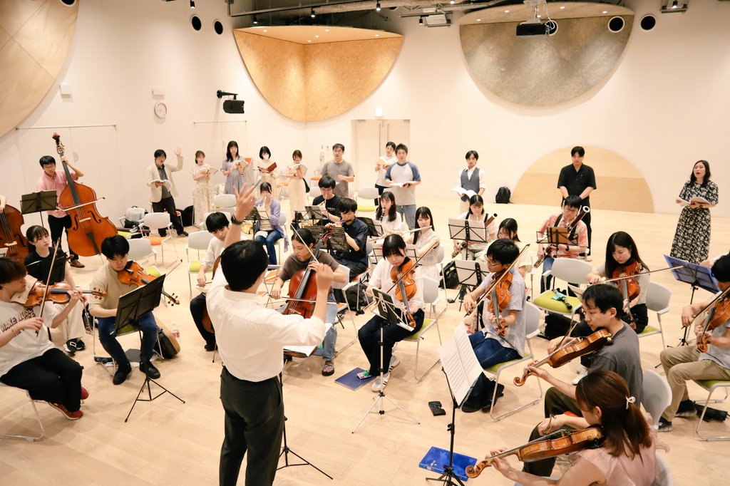 明るいホールの中で稽古に励むオーケストラ、歌手たちの写真