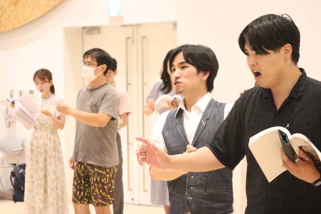 楽譜を片手に歌う歌手山本 和之と並ぶ白石渉そしてその後ろで歌っている歌手たち