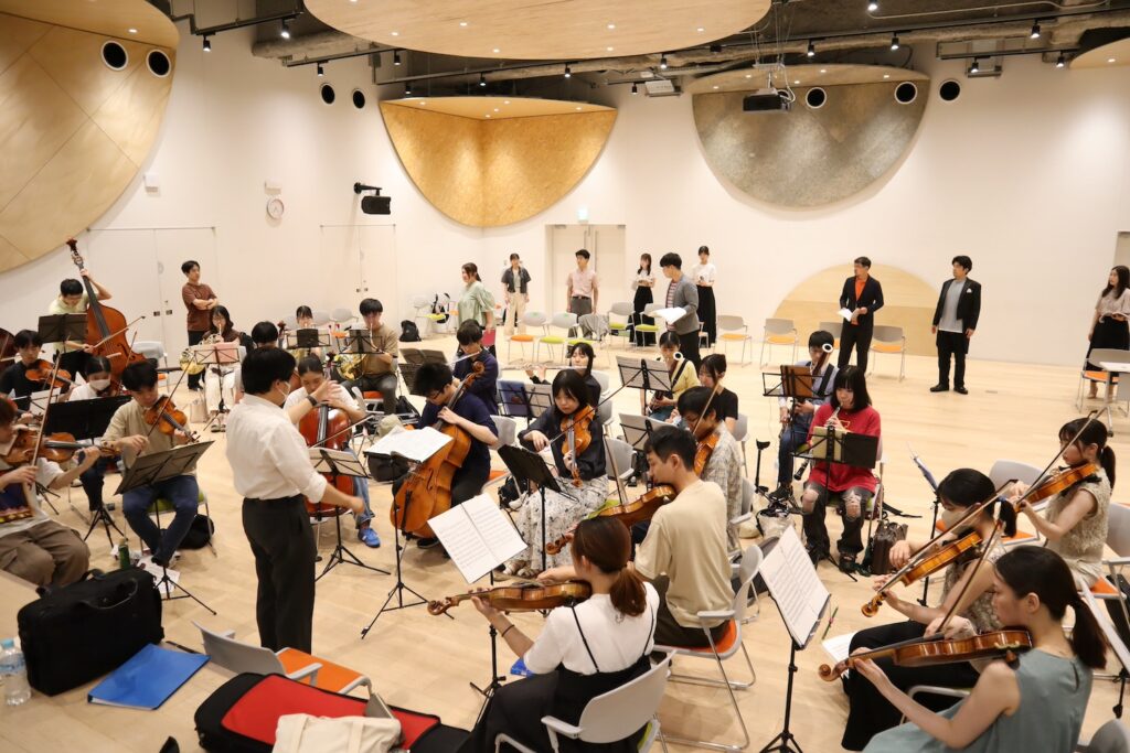 明るいホールの中で稽古に励むオーケストラ、歌手たちの写真